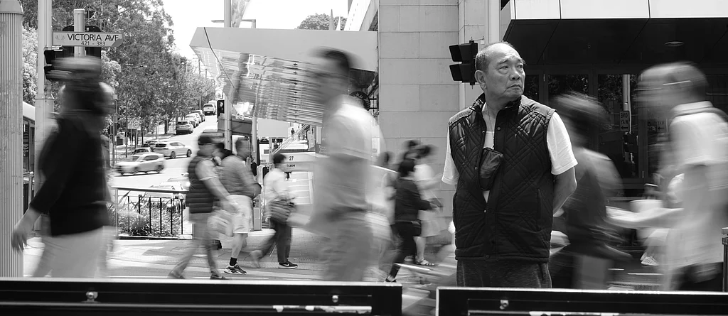 Black and white photo of a man standing still amidst a busy, blurred street scene.